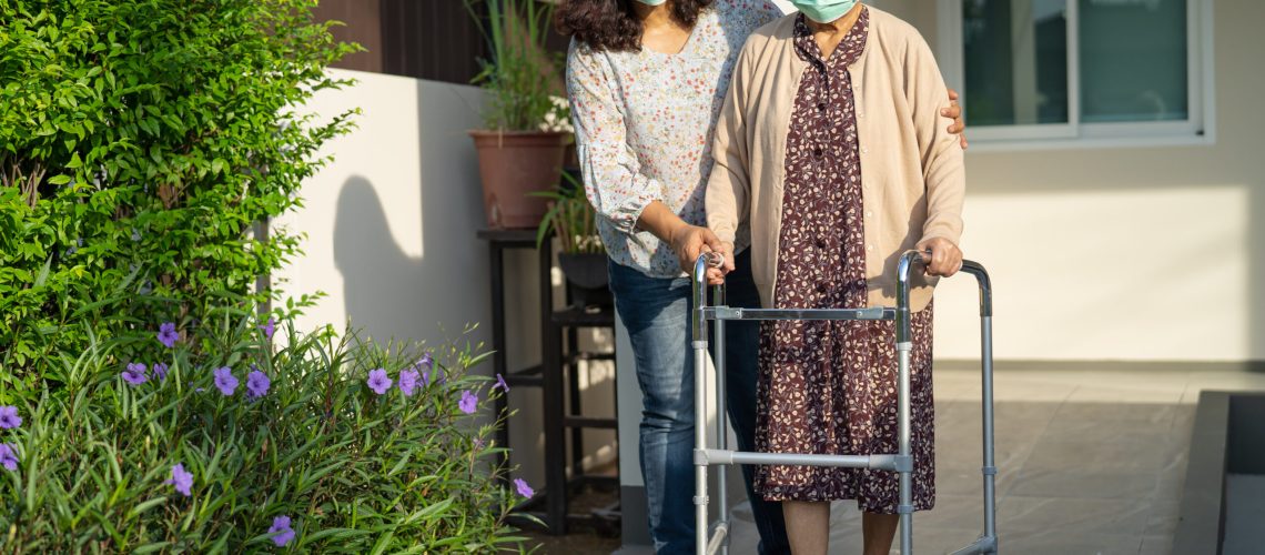 Caregiver help Asian senior or elderly old lady woman walk with walker on ramp and wearing a face mask for protect safety infection Covid-19 Coronavirus.